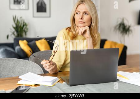 Discouraged middle-aged woman sitting at the laptop, does not know what to do at office, looking away, worried and tired, an entrepreneur woman is thinking how to save business in the crisis Stock Photo