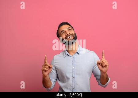 Cheerful overjoyed Indian guy points fingers up at empty copy space, isolated on pink background. Multiracial eastern man recommends deal, pays yours attention, presenting new product, advertising Stock Photo