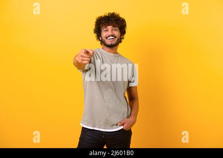 Smiling young indian man with curly hairstyle pointing finger at you standing isolated on yellow background, cheerful teenage guy indicating at the camera Stock Photo