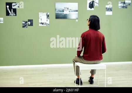Rear view of unrecognizable young man sitting on stool in modern art gallery looking at black and white photos on green wall Stock Photo