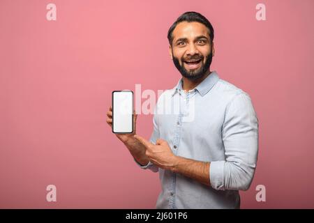 Surprised Indian man holds smartphone in a hand and points finger at empty phone screen, astonishment hispanic guy presenting best deal, advertising new mobile app, isolated on pink Stock Photo