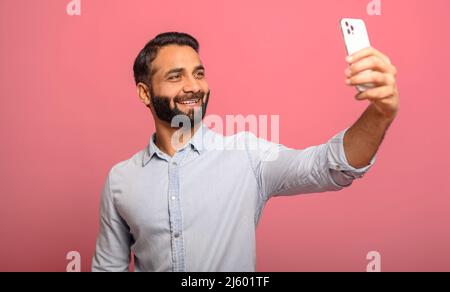 Photo of handsome cheerful smiling Indian man extends hand with smartphone, taking selfie portrait isolated over pink background. Hispanic guy photographs himself, streaming live in social media Stock Photo