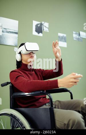 Young Asian man with disability wearing VR headset visiting contemporary photography exhibition in modern art gallery using augmented reality technolo Stock Photo