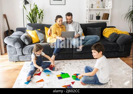 Parents working on the distance during quarantine. Father and mother work remotely with laptops sitting on the couch in living room, two sweet kids playing with toys on the foreground Stock Photo