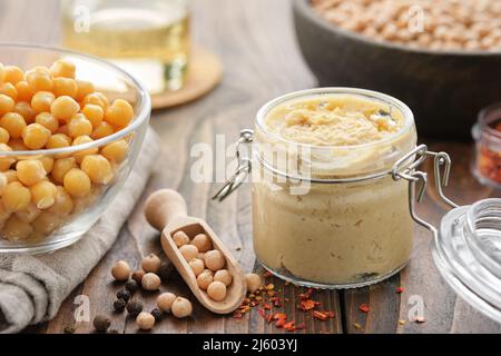 Jar of hummus sauce dip and chickpeas grains in bowl. Scoop of whole chick peas and spices on table. Stock Photo