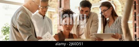 Analyzing results. Multicultural team holding reports and looking at the laptop while working together in the modern office Stock Photo