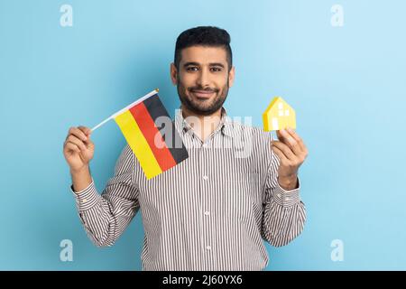 Satisfied man standing with paper house and flag of German, immigration and house purchase, rental housing, wearing striped shirt. Indoor studio shot isolated on blue background. Stock Photo