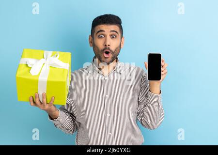 Amazed bearded businessman holding present box and smatrphone with blank screen for promotion, looking at camera with open mouth, wearing striped shirt. Indoor studio shot isolated on blue background. Stock Photo