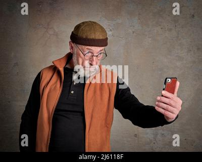 Head and shoulders portrait of a senior, bearded man taking selfie with his cell phone and making funny faces Stock Photo