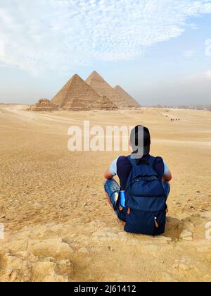 Egypt. Cairo - Giza. young blonde tourist girl with a backpack is sitting on the sand looking at General view of pyramids from the Giza Plateau. enjoy Stock Photo