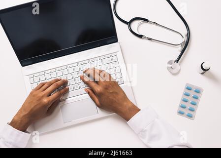 From above crop anonymous African American medical practitioner consulting remotely on netbook at table with stethoscope pills and bottle of syrup Stock Photo