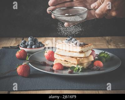 person powders freshly baked waffles with icing sugar using sieve or strainer, - homemade waffles are dusted with icing or powdered sugar and made rea Stock Photo