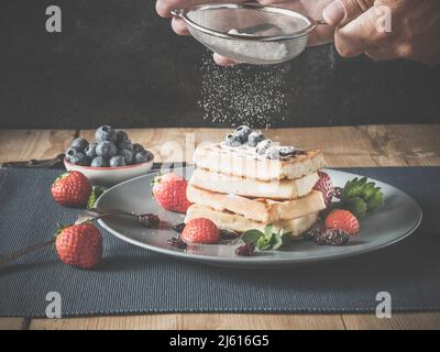 person powders freshly baked belgium waffles stack with icing sugar using sieve or strainer, - homemade waffles are dusted with icing or powdered suga Stock Photo