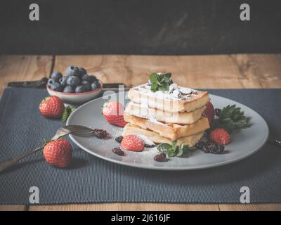 wooden table is decorated with a nice plate, placemate, fresh picked blueberries and strawberries delicious stack of fluffy waffles with blueberry top Stock Photo