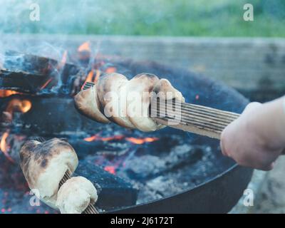 kids hand holding stick with stockbrot or twisted campfire stick bread over flames or outdoor campfire - family having a barbecue party in the garden Stock Photo
