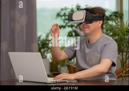 Asian man wearing VR headset and using laptop while sitting at his desk Stock Photo