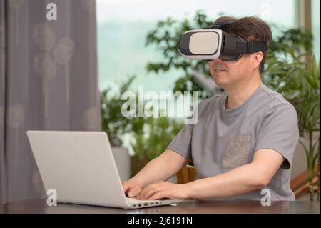 Asian man wearing VR headset and using laptop while sitting at his desk Stock Photo