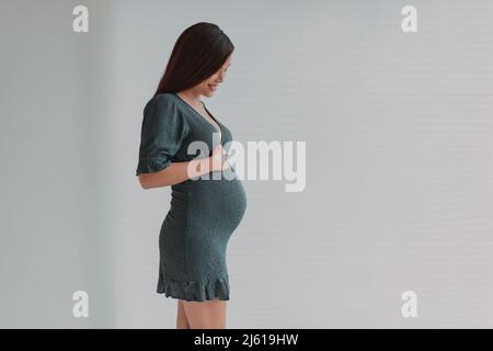 Beautiful pregnant young Asian woman looking down at her belly for maternity photoshoot wearing green dress. Pregnancy photos during third trimester Stock Photo