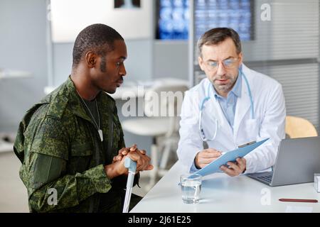 Mature doctor explaining checkup test results to young Black military man using walking stick after getting injured Stock Photo