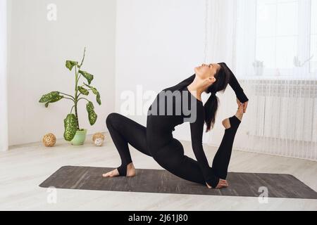 Woman in black sportswear practicing yoga doing a variation of the Eka Pada Rajakapotasana exercise, pigeon pose, exercising while sitting on a mat in Stock Photo
