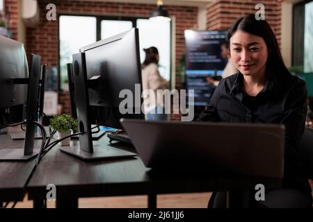Confident cyber security officer accesing administrator rights by bypassing firewall defence system. Asian cybernetics engineer writing lines of code in order to secure mainframe database storage. Stock Photo