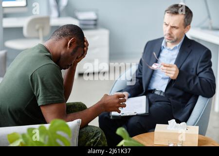 Depressed young adult Black soldier with PTSD remembering war at therapy session taking glass of water to drink Stock Photo