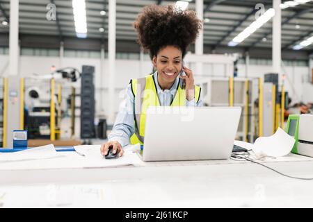 Smiling engineer working on laptop talking on mobile phone in factory Stock Photo