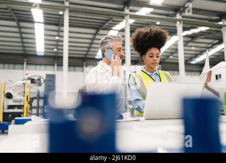 Technician with laptop standing by businessman talking on mobile phone in factory Stock Photo