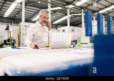 Businessman using laptop talking on smart phone in factory Stock Photo