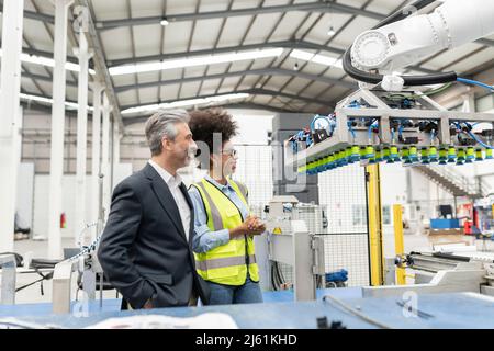Happy businessman with hands in pockets standing by engineer looking at robotic machine in factory Stock Photo