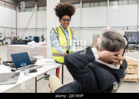Smiling engineer looking at businessman sitting with hands behind head in factory Stock Photo