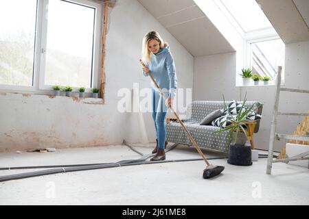 Smiling blond woman sweeping floor with broom in attic Stock Photo