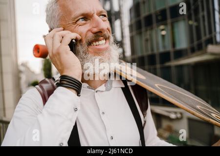 Grey mature man talking on cellphone while standing with skateboard at city street Stock Photo
