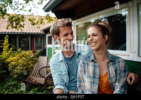 Smiling man looking at happy girlfriend sitting at backyard Stock Photo