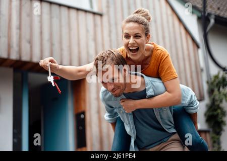 Smiling man giving piggyback ride to cheerful woman holding house key at backyard Stock Photo