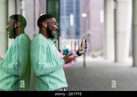 Happy man with wireless in-ear headphones having video call on smart phone by glass wall Stock Photo