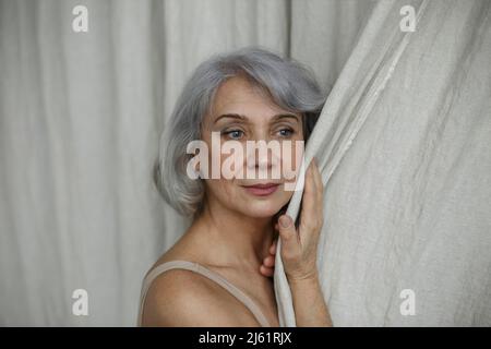 Thoughtful senior woman with gray hair touching curtain at home Stock Photo