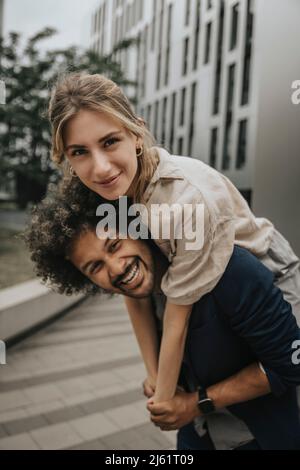 Happy young man giving piggyback ride to girlfriend in front of modern building Stock Photo