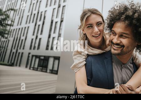 Smiling young man giving piggyback ride to girlfriend in front of modern building Stock Photo
