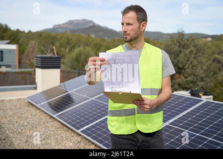 Engineer with blueprint by solar panels standing on rooftop Stock Photo