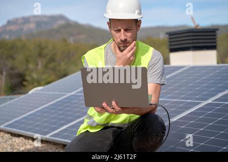 Engineer with hand on chin using laptop in front of solar panels Stock Photo