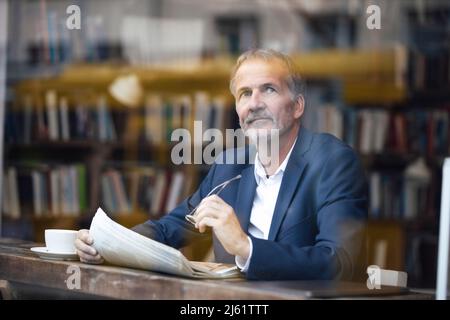 Thoughtful businessman with eyeglasses and newspaper sitting at table in cafe Stock Photo
