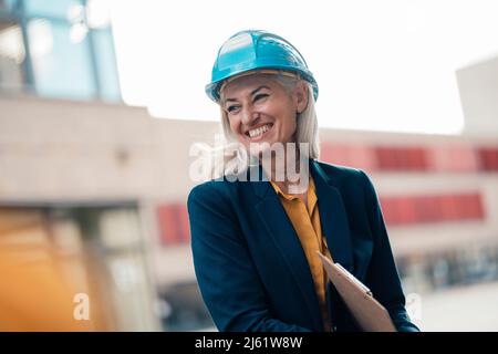 Cheerful female architect with clipboard wearing hardhat at office park Stock Photo