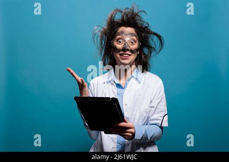 Crazy scientist with dirty face and messy hairstyle having touchscreen tablet acting shocked and mad after failed experiment explosion. Wacky looking chemist being baffled after lab discovery. Stock Photo