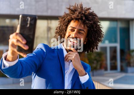 Happy businessman with hand on chin taking selfie through mobile phone in front of building Stock Photo