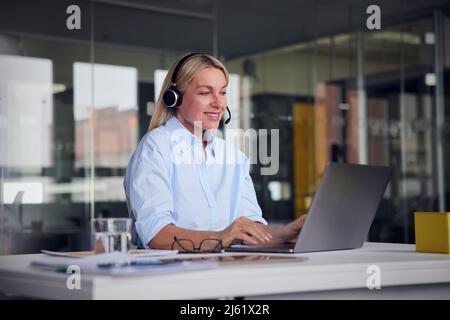 Smiling businesswoman wearing headset using laptop at desk in office Stock Photo