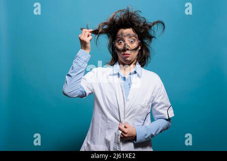 Wacky and crazy looking mad scientist grabbing messy hair while having dirty face from explosion on blue background. Insane and funny silly chemist acting dizzy after failed chemical experiment. Stock Photo