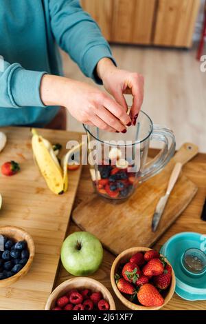Woman putting fresh chopped fruits in blender at table in kitchen Stock Photo