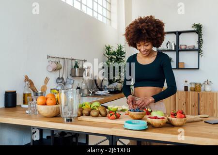 Smiling young woman cutting fruits standing at table in kitchen Stock Photo