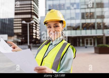 Happy female architect wearing hardhat holding blueprint Stock Photo